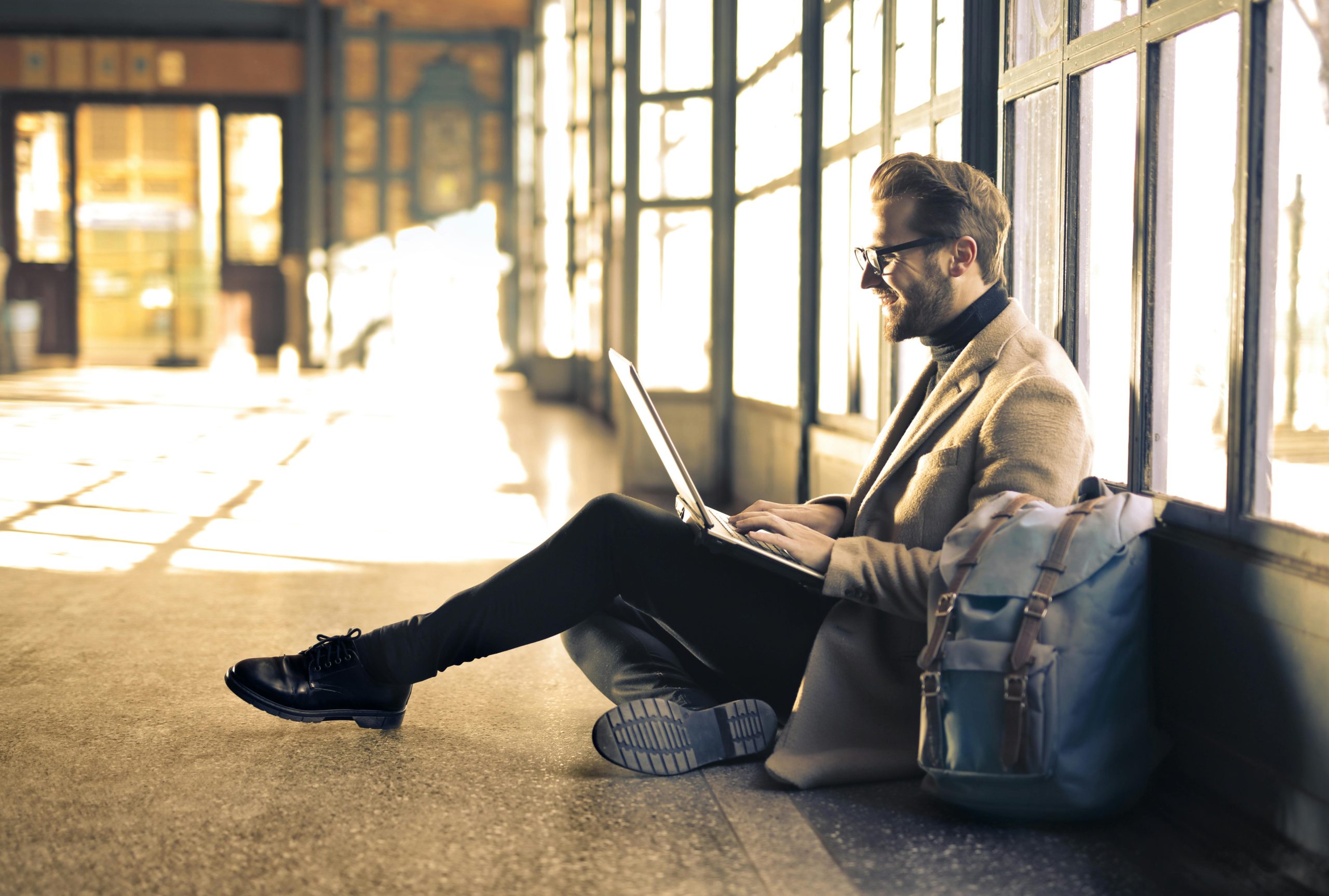 The featured image consists of a younger adult man sitting on the ground with his back against some windows on his laptop while smiling. He is wearing a nice fall jacket, black jeans, black shoes, and wearing studious black glasses. Next to his left side is a backpack.