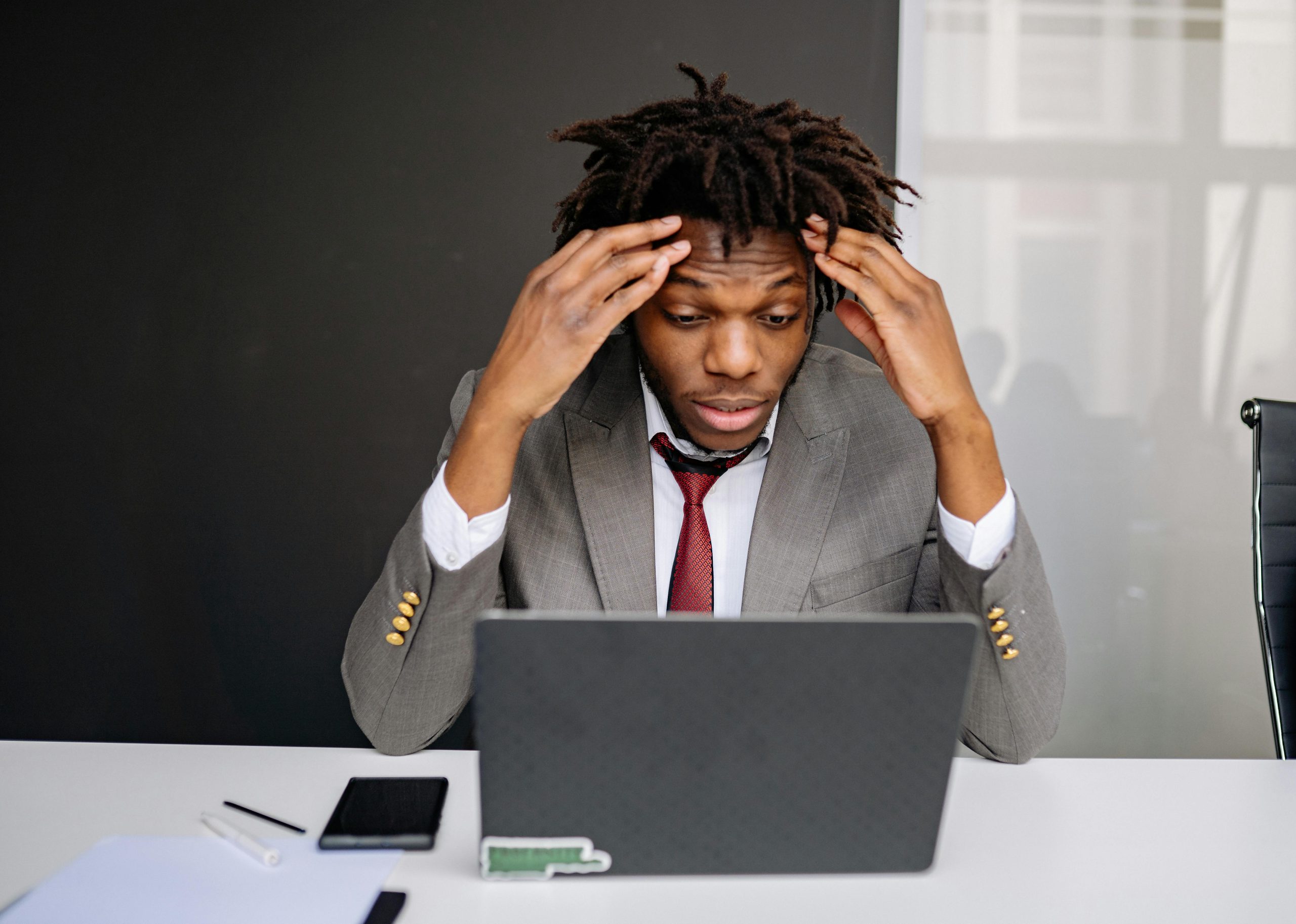 Distressed individual in a gray dress suit with his hands on his forehead looking at his laptop. The individual could possibly be looking at a fake review about his product or brand on his computer.