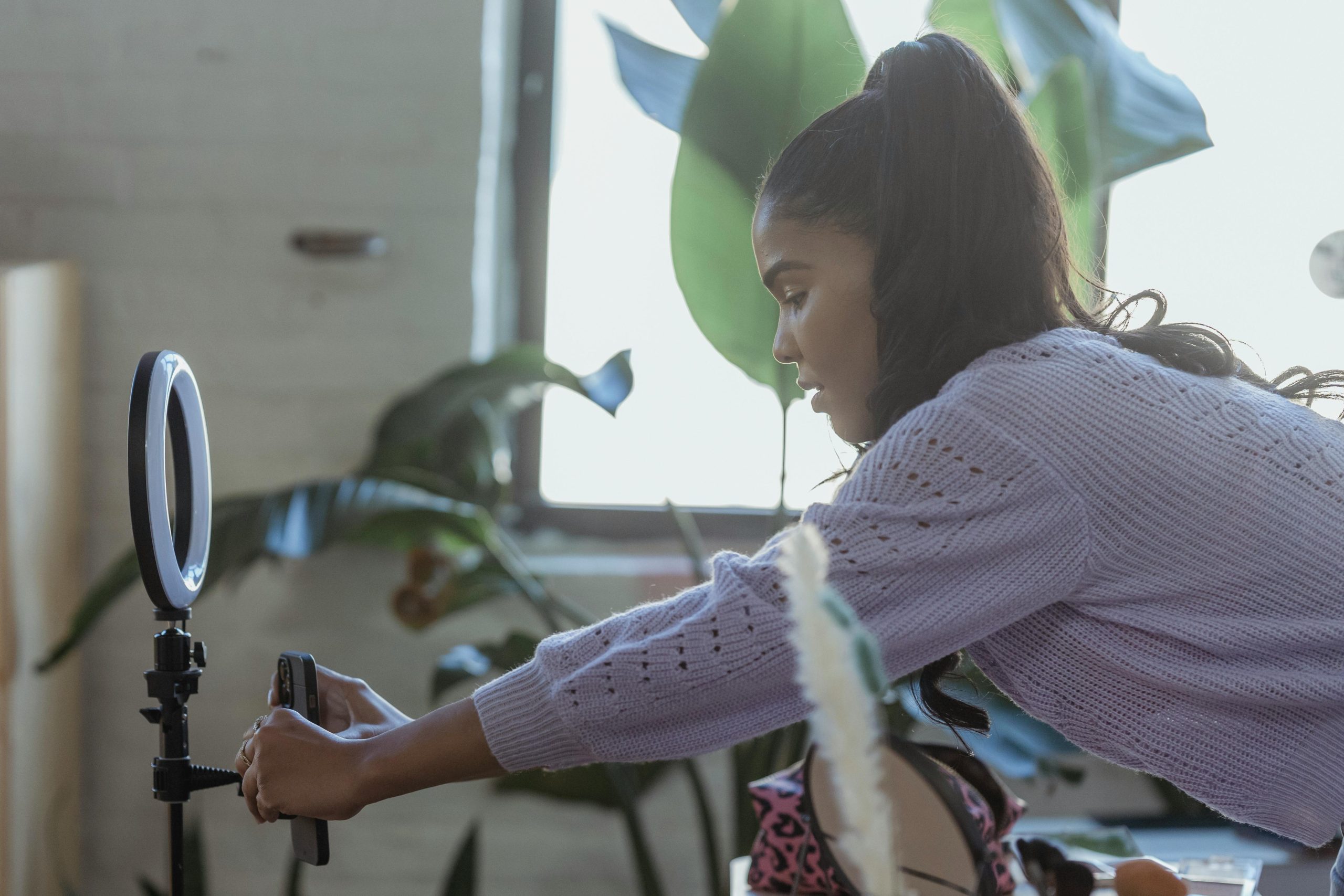 The featured image consists of a girl with a black/brown ponytail leaning over and adjusting her phone on a tripod that has a white ring light on the top of it. The girl is wearing a light pink sweater with decorative holes in it. Additionally, there is a window in the background with large green leafs in front of it.