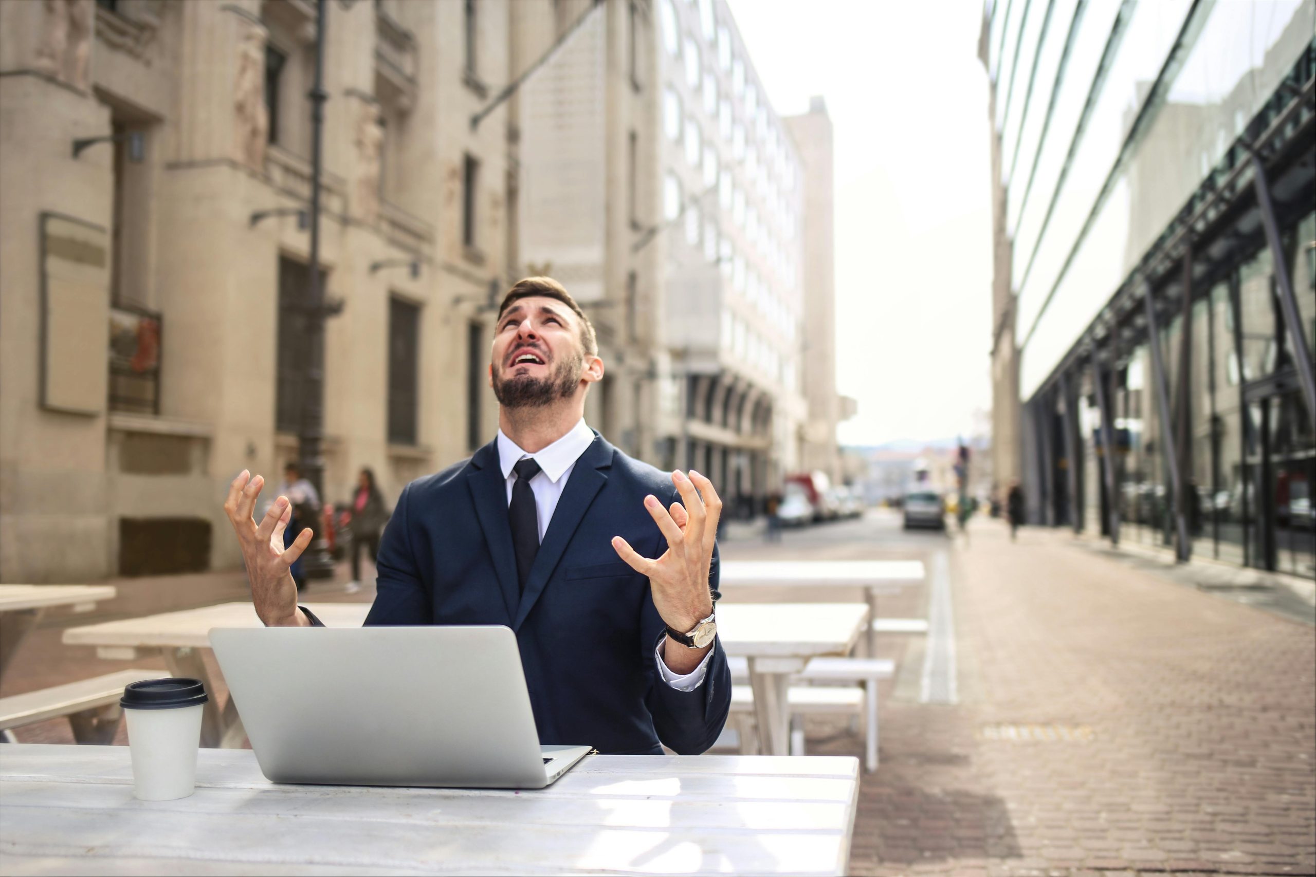 The featured image showcases a man in a business suit sitting at an outside table looking up at the sky with his hands held up in frustration. In front of him on the table is his laptop and coffee. He appears to be on a brick road with stone buildings in the background.