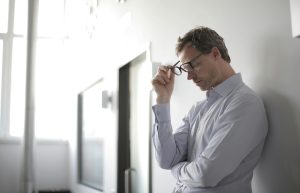 Picture of a disappointed-looking man holding his glasses, looking down, with his back against the wall.