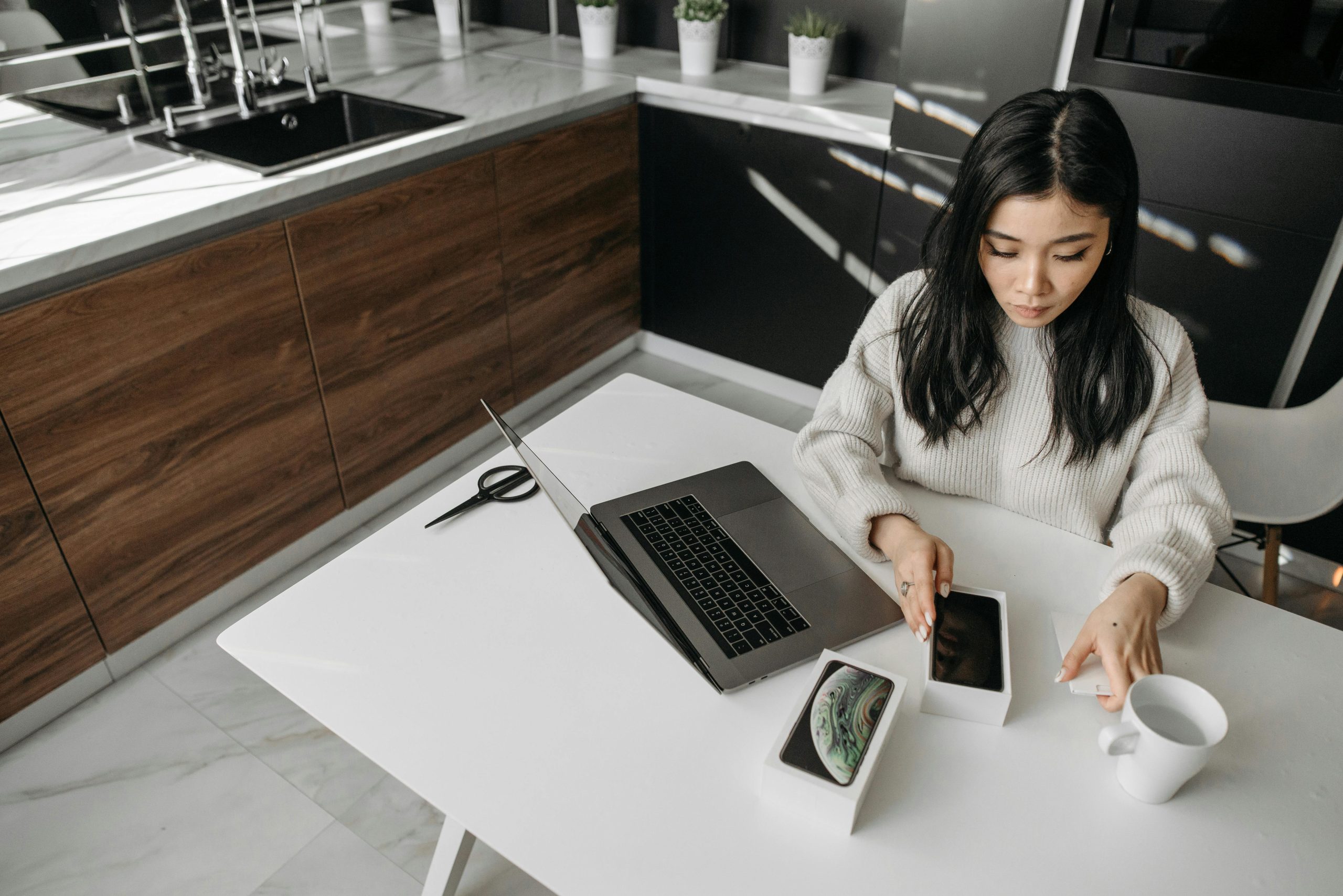 The featured image showcases a woman sitting at her kitchen table opening a brand-new i-Phone. Next to her is her opened laptop, a pair of scissors, and a coffee mug.