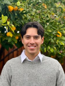 Young man with dark hair is standing in front of some greenery.