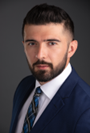 Image of a man with dark hair and a beard dressed in a dark suit with a white shirt and tie.