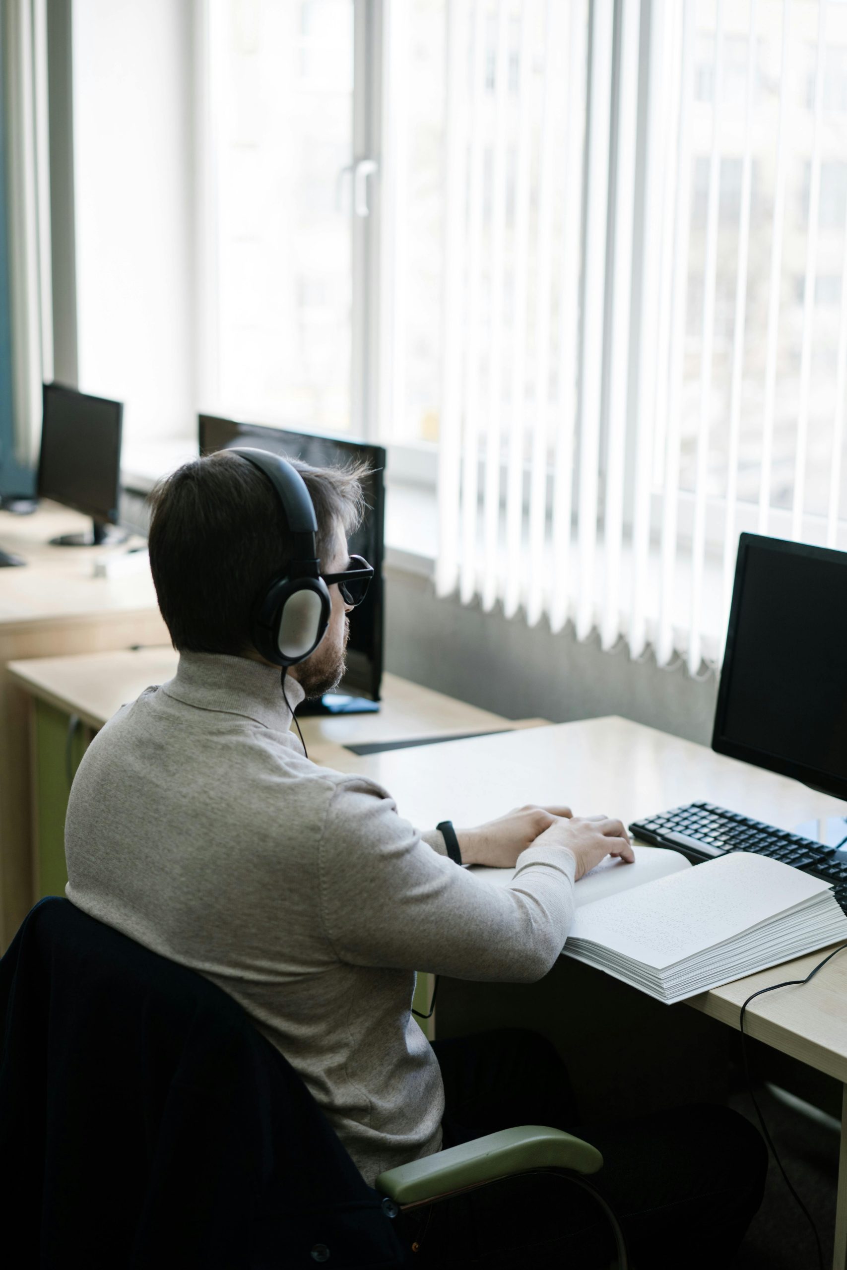 A blind man wearing headphones sits in front of a computer while reading a braille book.