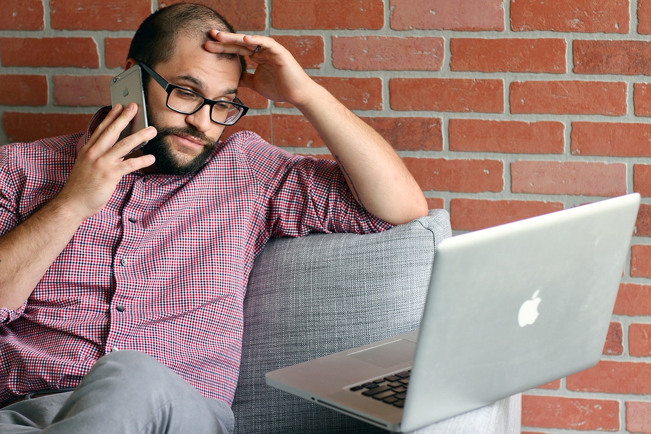 A man sits on a couch, looking very stressed, with a phone to his ear while he stares frustratingly at his computer screen.