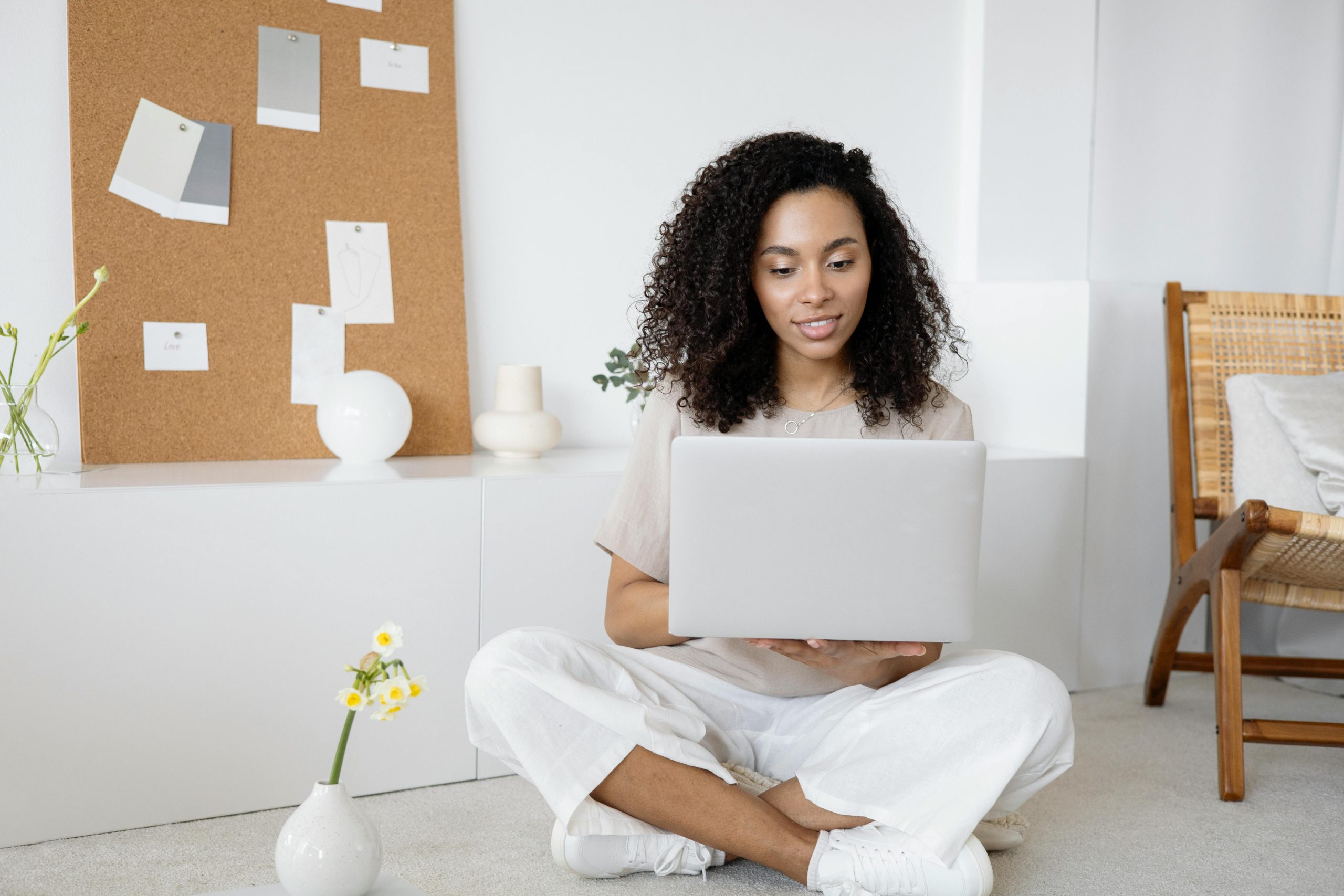 The image shows a girl with curly hair sitting cross-legged on a white carpeted floor who is wearing white and holding her laptop in her hands. Beside her are white cabinets with decorations on it and a bulletin board. On the ground, there is a small pot of white and yellow flowers next to her and a brown and tan wooden/wicker chair in the background off to the side with a white pillow on it.