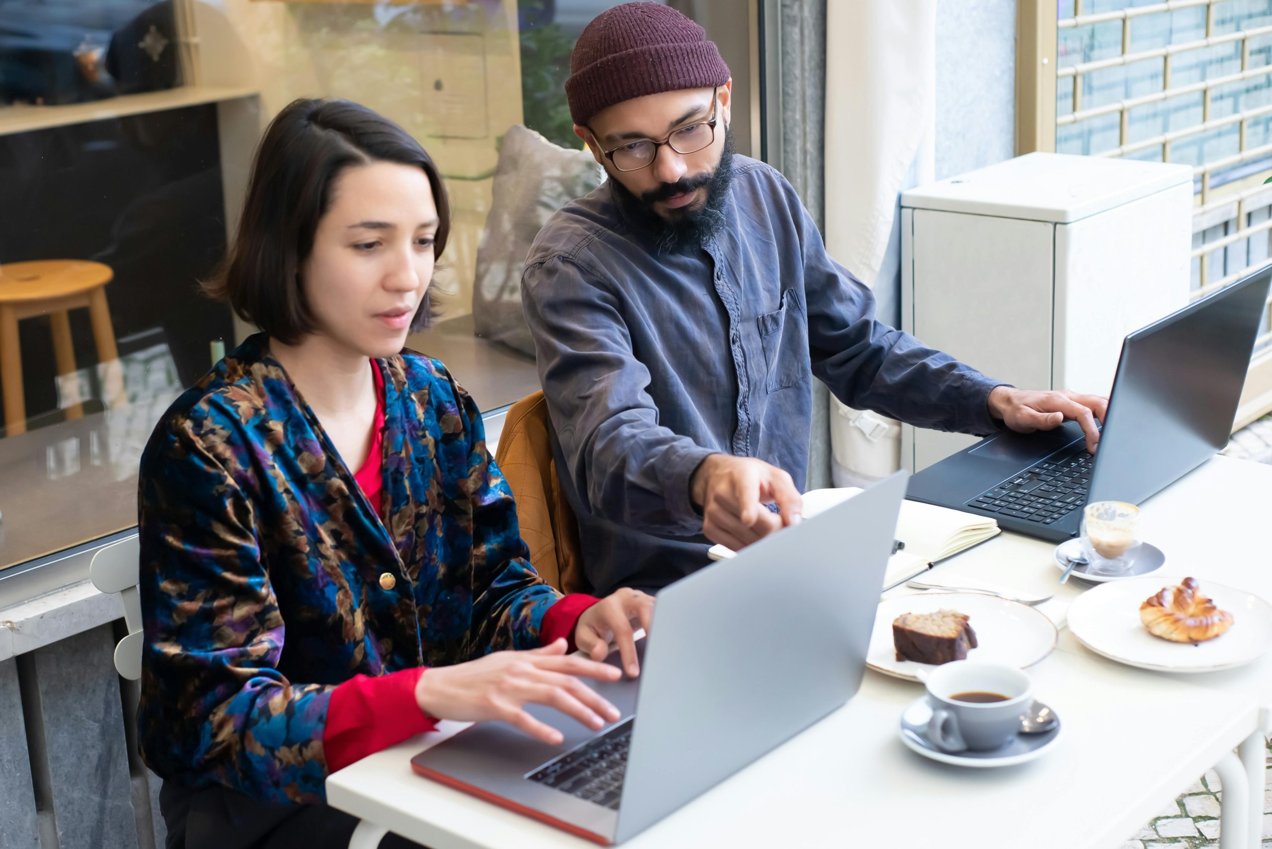 The featured images showcases a studios-looking man with a maroon beanie, black and gray beard, glasses, and a long-sleeve button-up navy blue shirt pointing at a girl's laptop screen. The girl, who is sitting to the man's right, is wearing a multi-colored silky cardigan with a long-sleeve red shirt underneath. The two are in a cafe with two sweet breads on white round plates and two coffees on a white table. There is a glass window behind the two individuals showing another room that has a wooden stool in it.