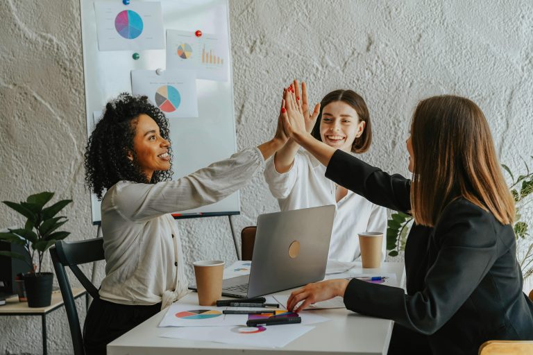 The featured image showcases three women dressed in casual office attire high-fiving each other at a table in the office. On the table, there are coffee cups, a laptop, pens, and pie charts. Behind them is a medium-sized whiteboard with additional pie and bar charts stuck onto it. There are also medium-sized and small plants near the women. Behind them is a white stone wall.