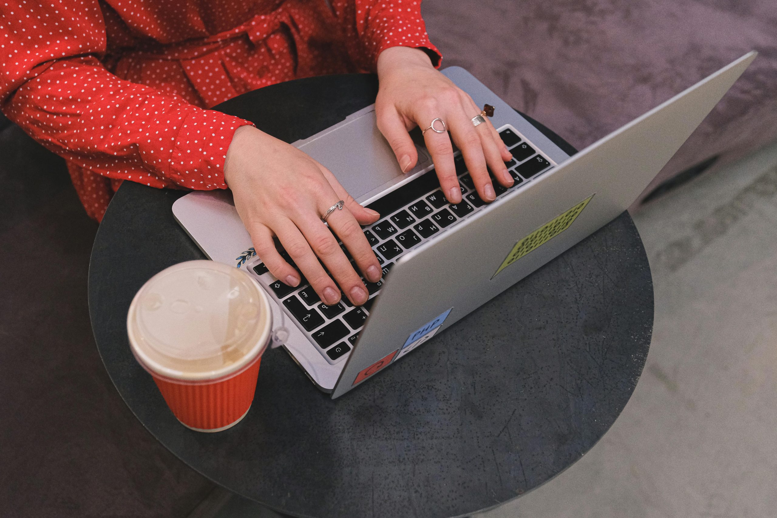 The picture shows a lady the hands of a lady in a red shirt with small white polkadots typing on her laptop, which is on a small round black table with a cup of coffee next to the laptop on the table.