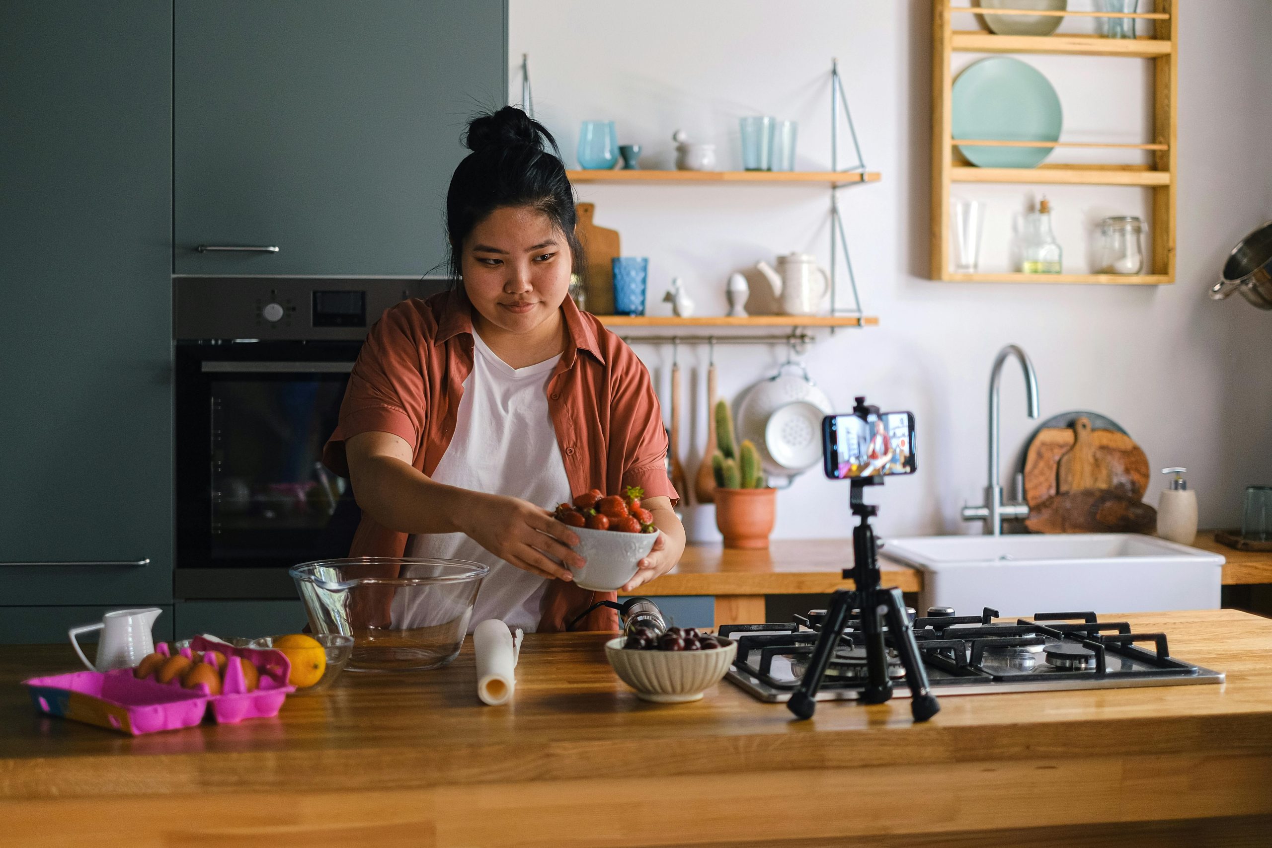 A girl wearing an apron holding a bowl of strawberries. In front of her is a kitchen counter, and on the counter, there is a carton of eggs to her left, and a mini tripod holding up her phone, which is filming her.