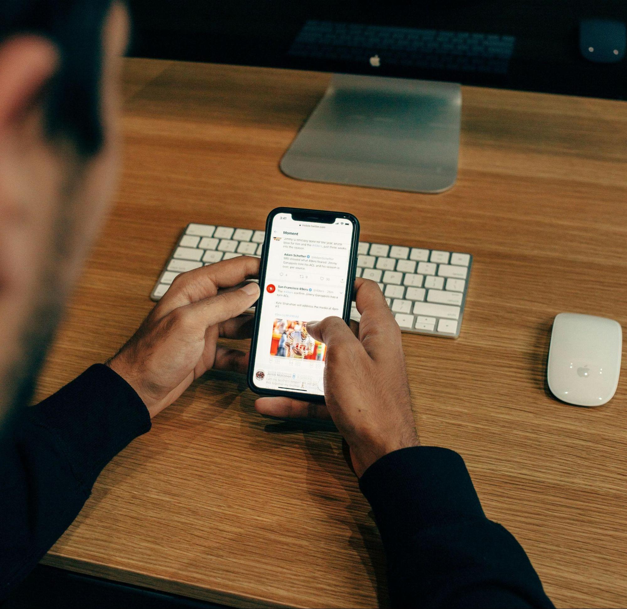 The featured image showcases someone's hands resting on a wooden table holding their smartphone. The person is scrolling through their Twitter feed with their thumb. Behind their phone is a wireless keyboard with white keys and a white, wireless mouse to their right.