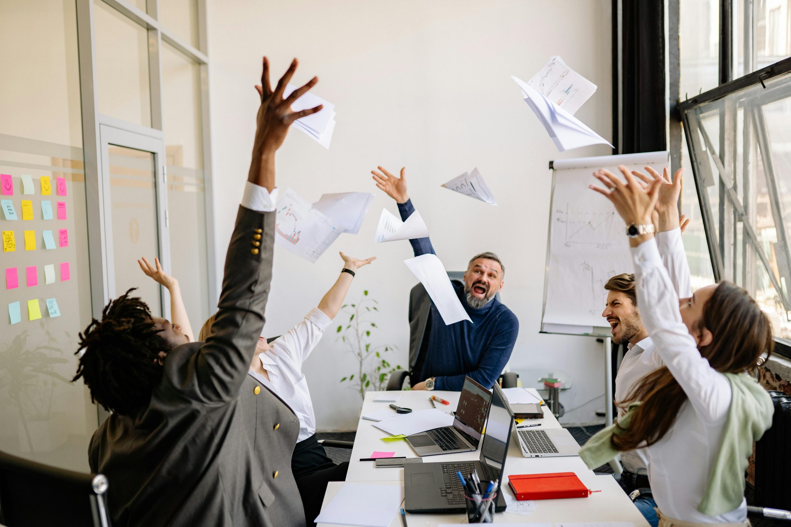 The featured image consists of 5 individuals in business attire throwing papers up in the air. On the table in front of them, there are laptops, notebooks, pens, sticky notes, and markers. On the left side of the image is are multiple glass windows and a glass door with sticky notes on it. To the right is a window that is open. In the back right corner is a large gray/silver stand holding up large sheets of paper that you can right on to illustrate ideas.