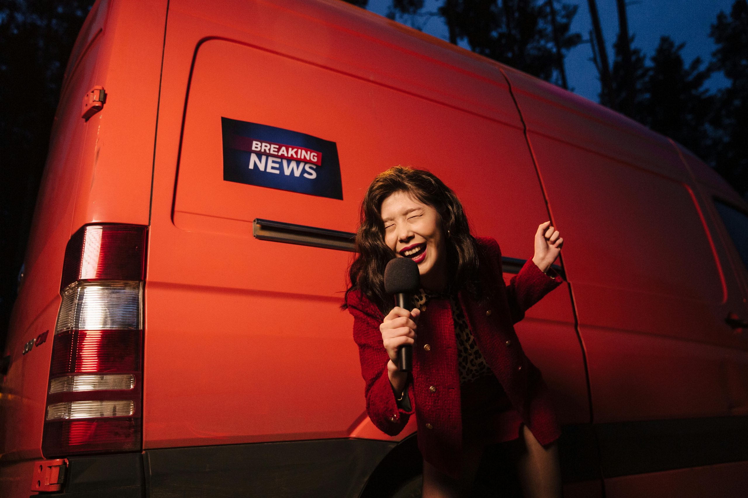 The featured image consists of a woman standing in front of a orange van that has a screen reading, "BREAKING NEWS" on it. The woman is holding a microphone and is wearing a velvety-red overcoat. She is outside and it is night time.