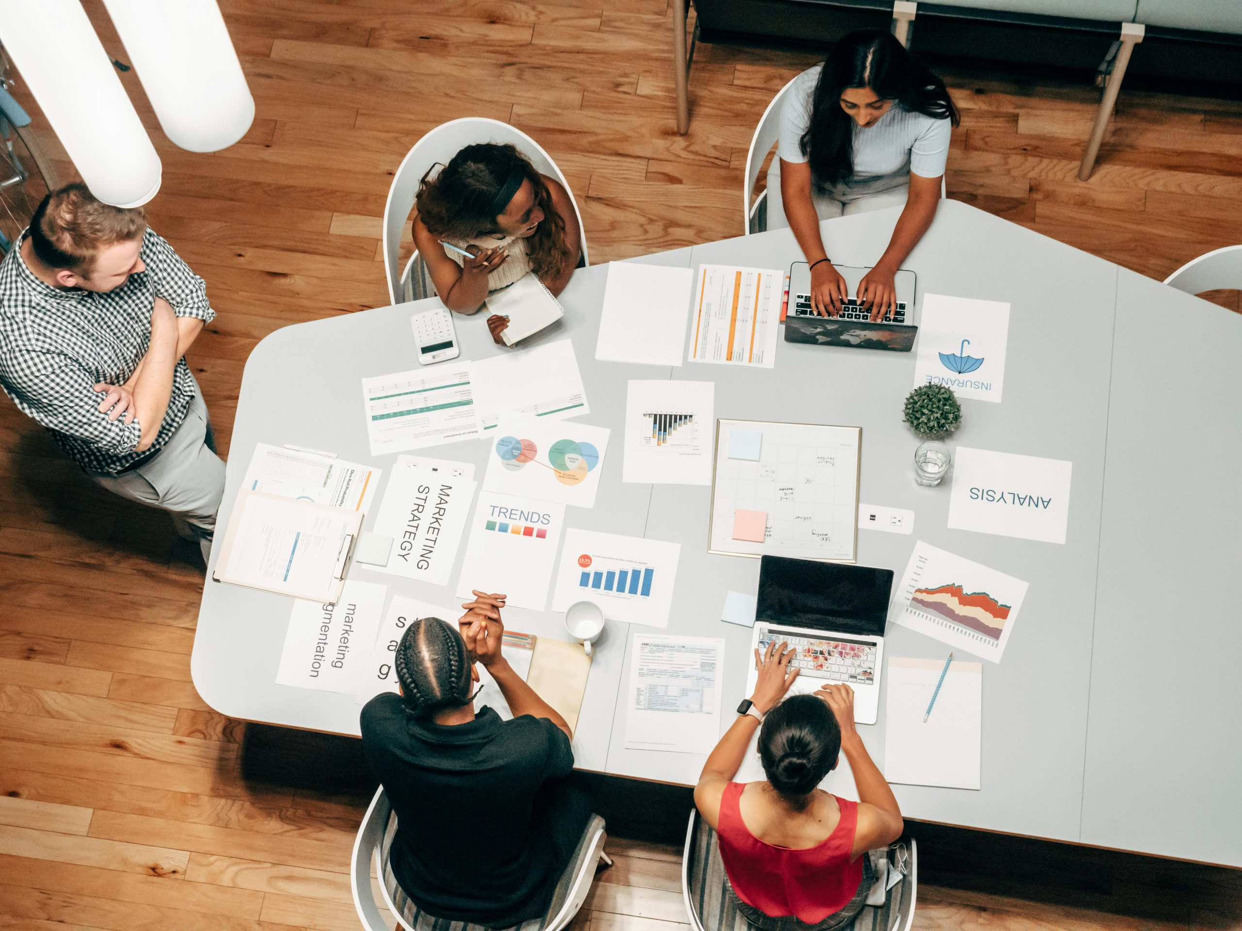 The featured image consists of an above snapshot of a marketing team of 5. They are seated around a gray table with papers that read "trends", "analysis", and "marketing strategy", alongside their laptops, pencils, and their notes.