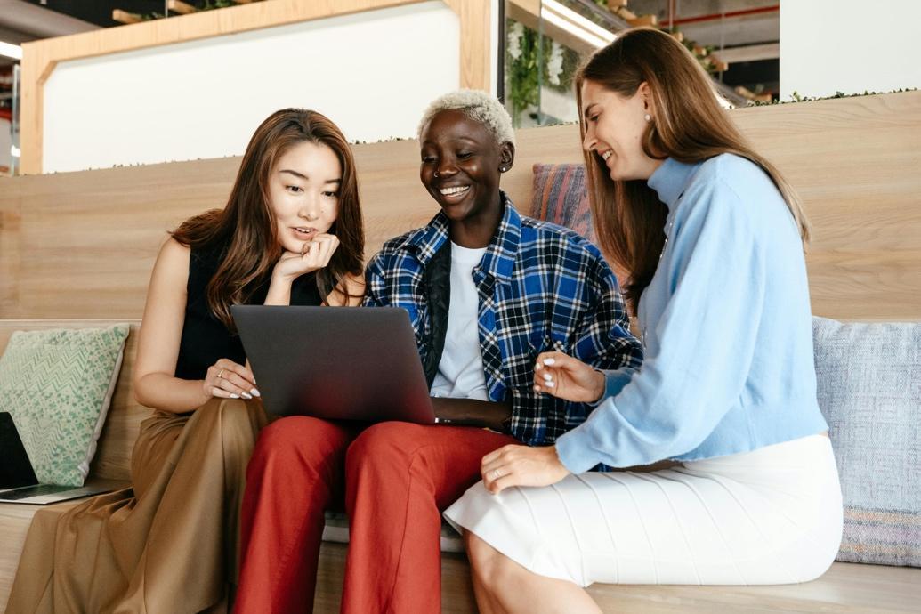 The featured image showcases a group of diverse women sitting down looking at a dark gray laptop in the women who is sitting in the middle's lap. They are all smiling. They are seated on a maple or ash bench with light blue and light green, ornate pillows next to them. It appears they are located in a modern office setting.