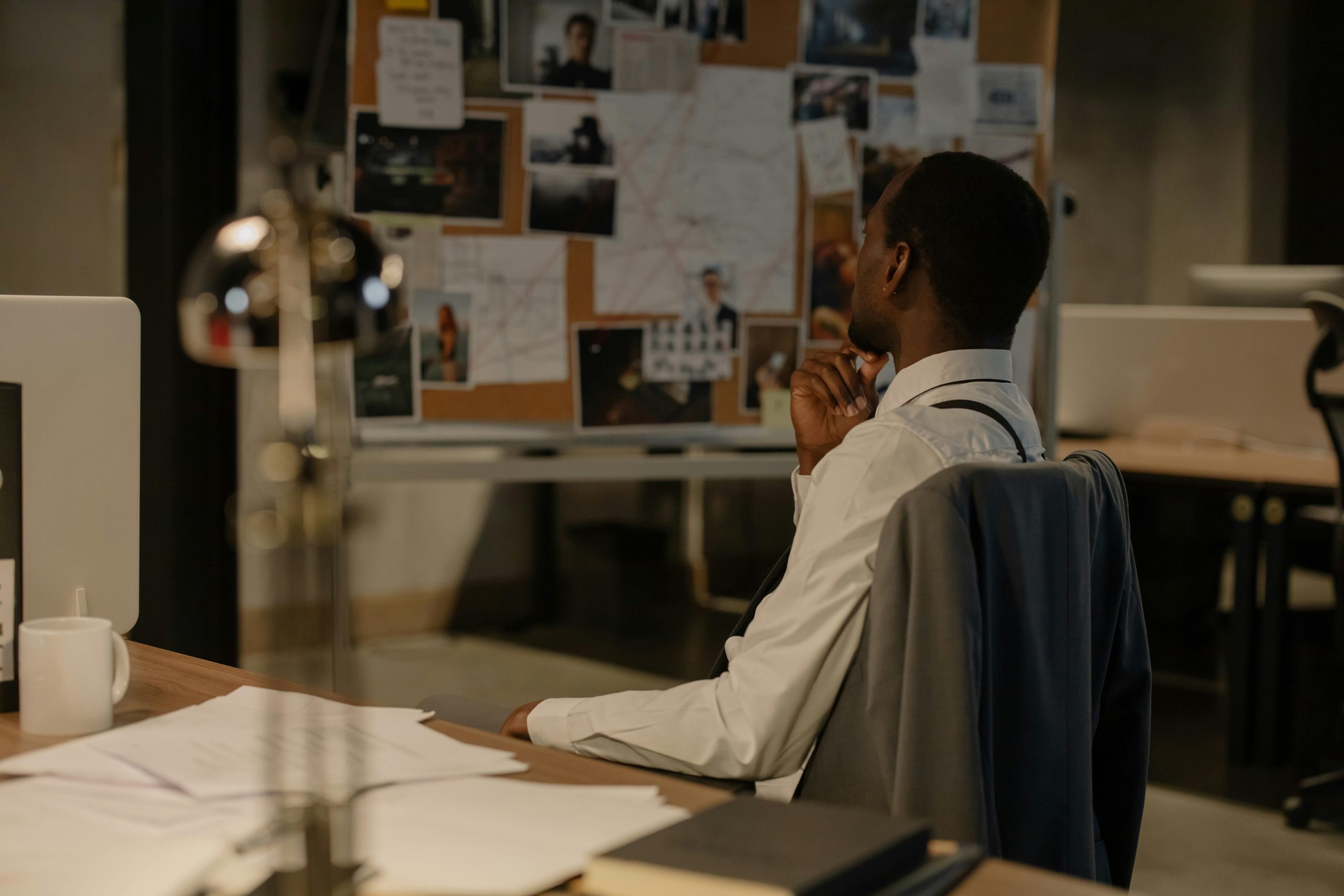 The featured images showcases a man in his computer chair at a desk with papers, a coffee cup, a book, and a lamp. He is looking across the room at a cork/bulletin board with papers and pictures with strings and thumbtacks. The man appears to be a detective.