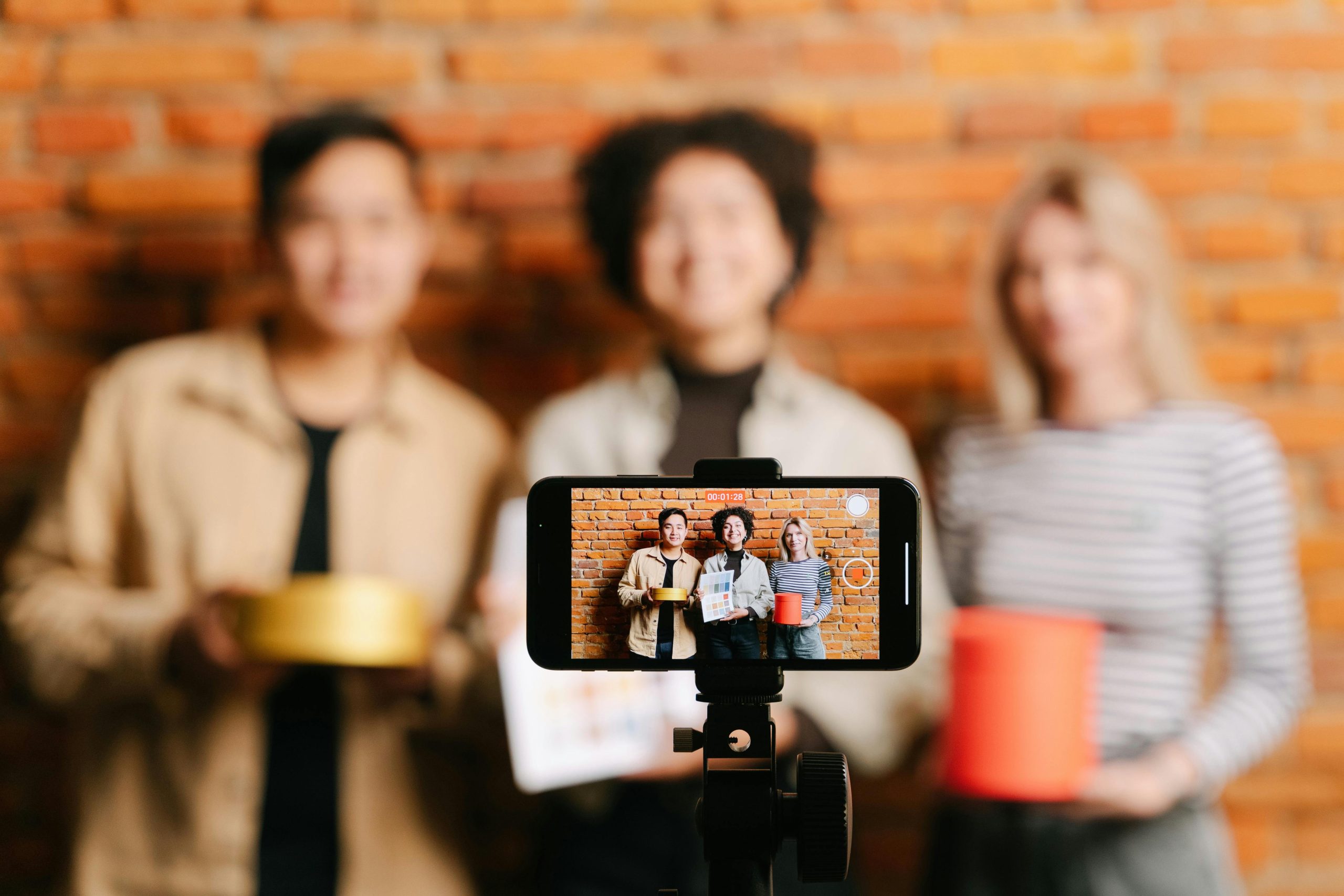 The featured image consists of a group of blurred-out people standing with their backs against a red brick wall. In front of them is a smartphone turned horizontally on a phone camera stand. On the phone, we can see that it is recording a video, and the people in the frame are holding a gold and red cylinder, both different sizes, and the person standing in the middle is holding up a piece of paper that shows multiple different shades of colors.