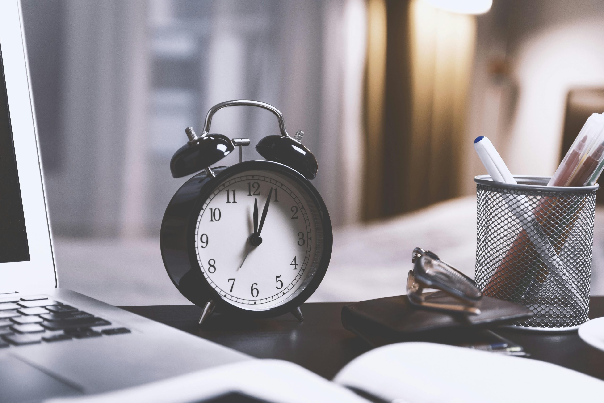 The featured image consists of a black, metal alarm clock on a black table with a grated cup holding pens and markers on the right, and a gray laptop to the clock's left.