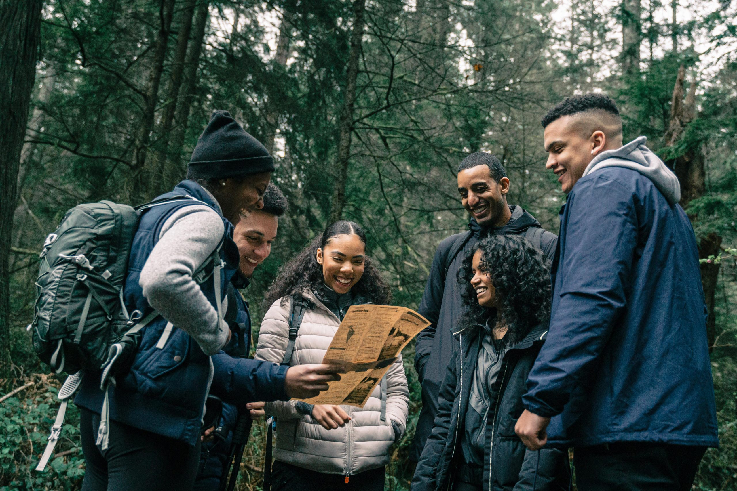 The featured image consists of a group of young hikers, men and women, looking at a map and smiling. They are wearing coats, and are in a the woods, where they are surrounded by green pine trees.