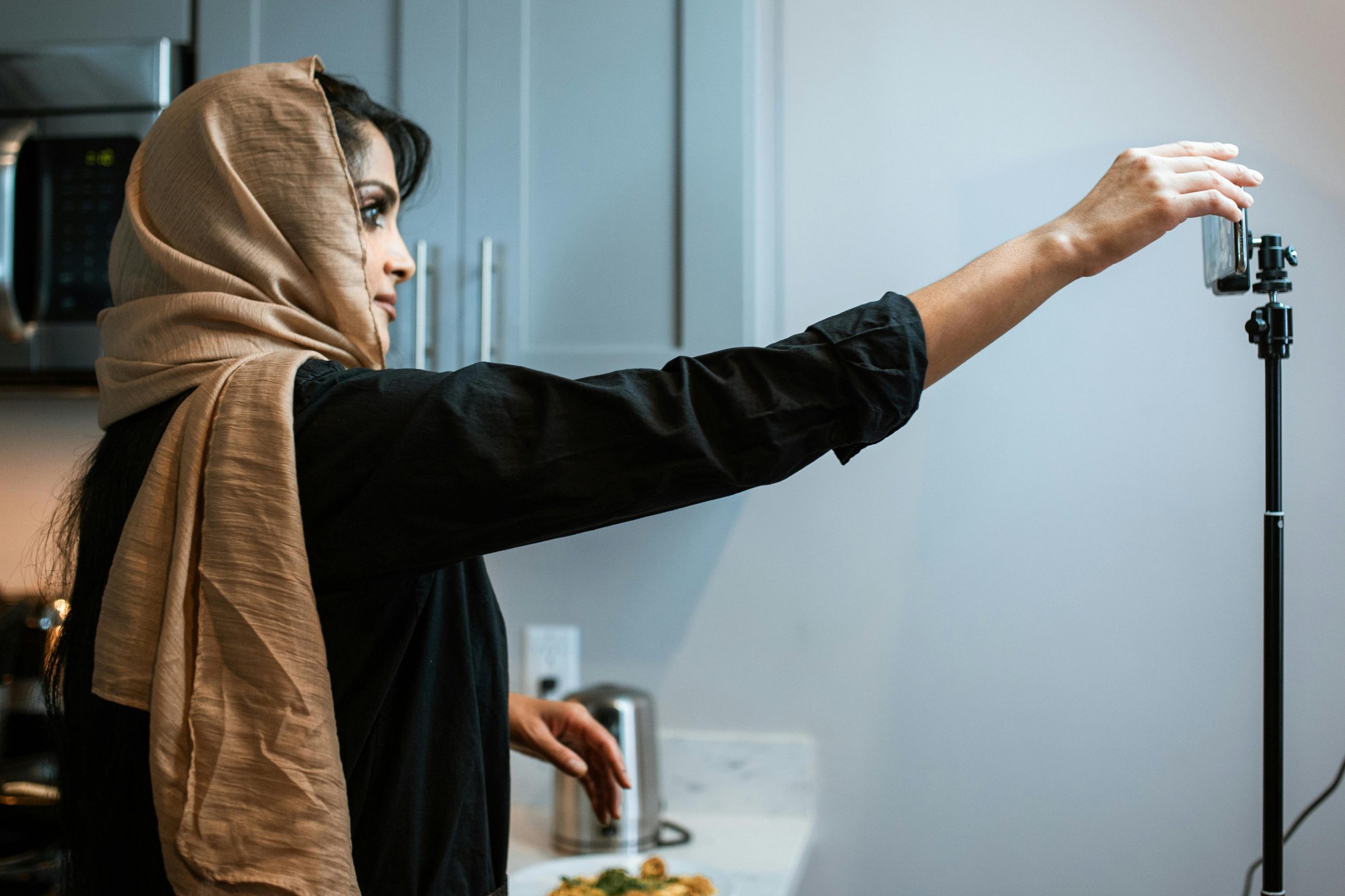 The featured image consists of a woman filming touching her smartphone, which is on a smartphone camera stand, in a kitchen.