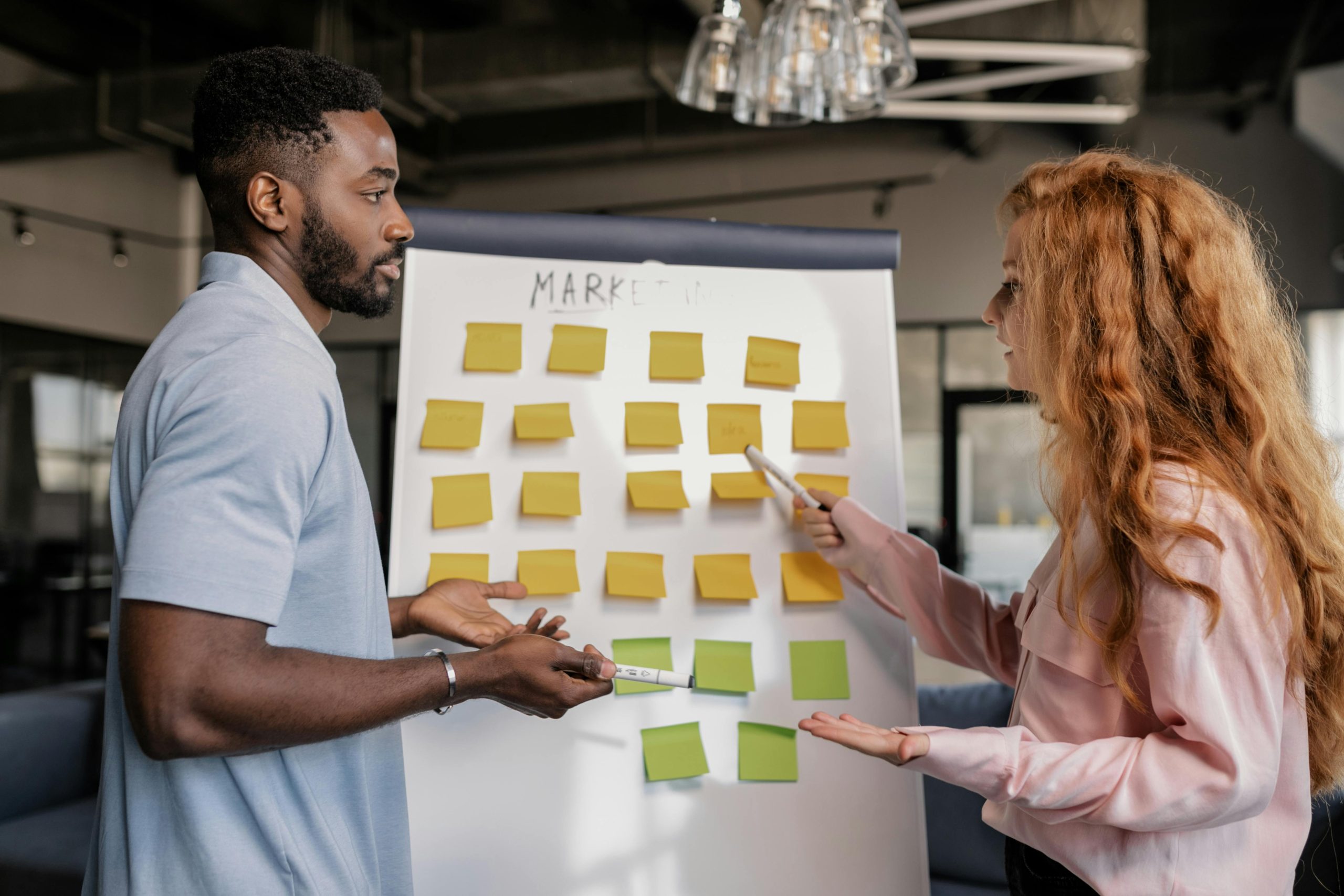 The featured image showcases a man and woman talking in front of a whiteboard on its easel that reads "marketing." There are several orange and green sticky / post-it notes on the white board. The two appear to be in a modern office setting.