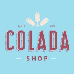 Colada Shop's company logo consists of a light-blue background with the dusty red, capitalized words "COLADA SHOP" in the center. The word shop is underneath the word Colada, and is in a smaller font size. Above the word colada are three white feathers (one pointing straight up, with the other two pointing to the right and left corners of the image respectively. On the left side of the feathers is the dusty red, capitalized text "CAFE" and to the right of the feathers is the capitalized, dusty red text "BAR"
