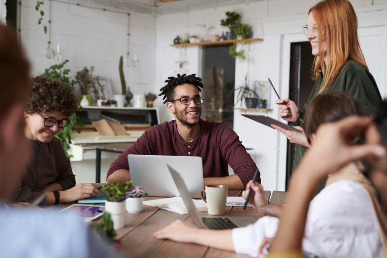 The featured image features a group of 6 young professionals sitting at a brown wooden table smiling at each other. There are a couple of open laptops, notebooks, plants, and a coffee cup on the table. A girl who is standing to the right is holding a smart tablet with a stylus. They appear to be in a modern-looking office with white brick walls and light-colored shelves holding various green plants.