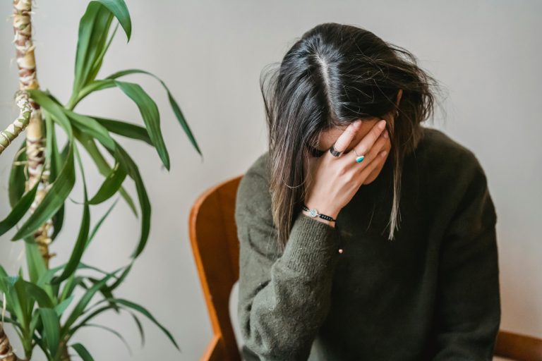 The blog's featured image showcases a woman sitting in a chair with her head in her right palm. She is clearly distressed about something. She has black hair with blond highlights/tips. She is wearing a forest-green sweater and has rings and a bracelet on. She is sitting in a wooden chair with a small tree with green leafs to her right. The wall behind her is white.
